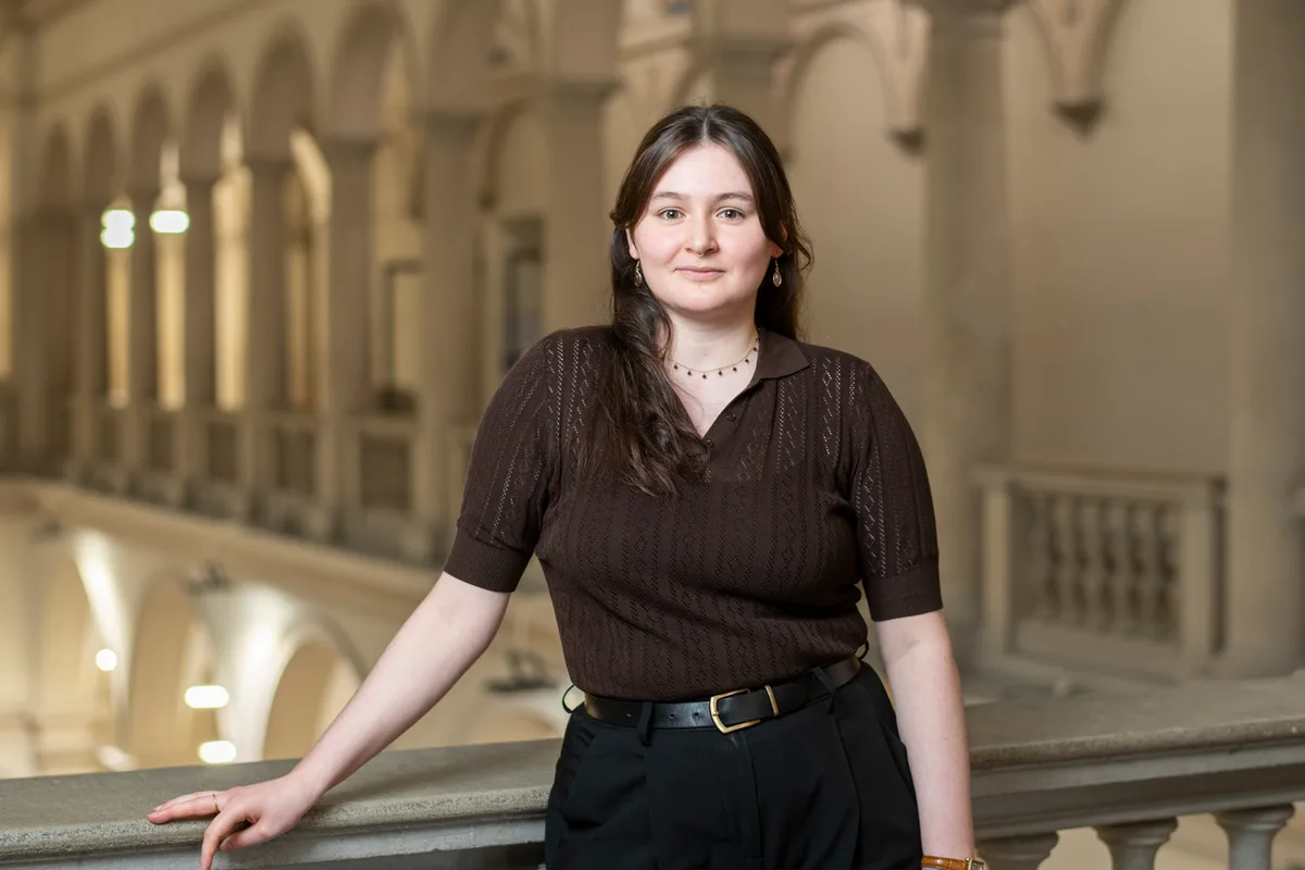 Porträt einer jungen Frau. Sie lehnt an ein Geländer im Innenhof einer Universität.